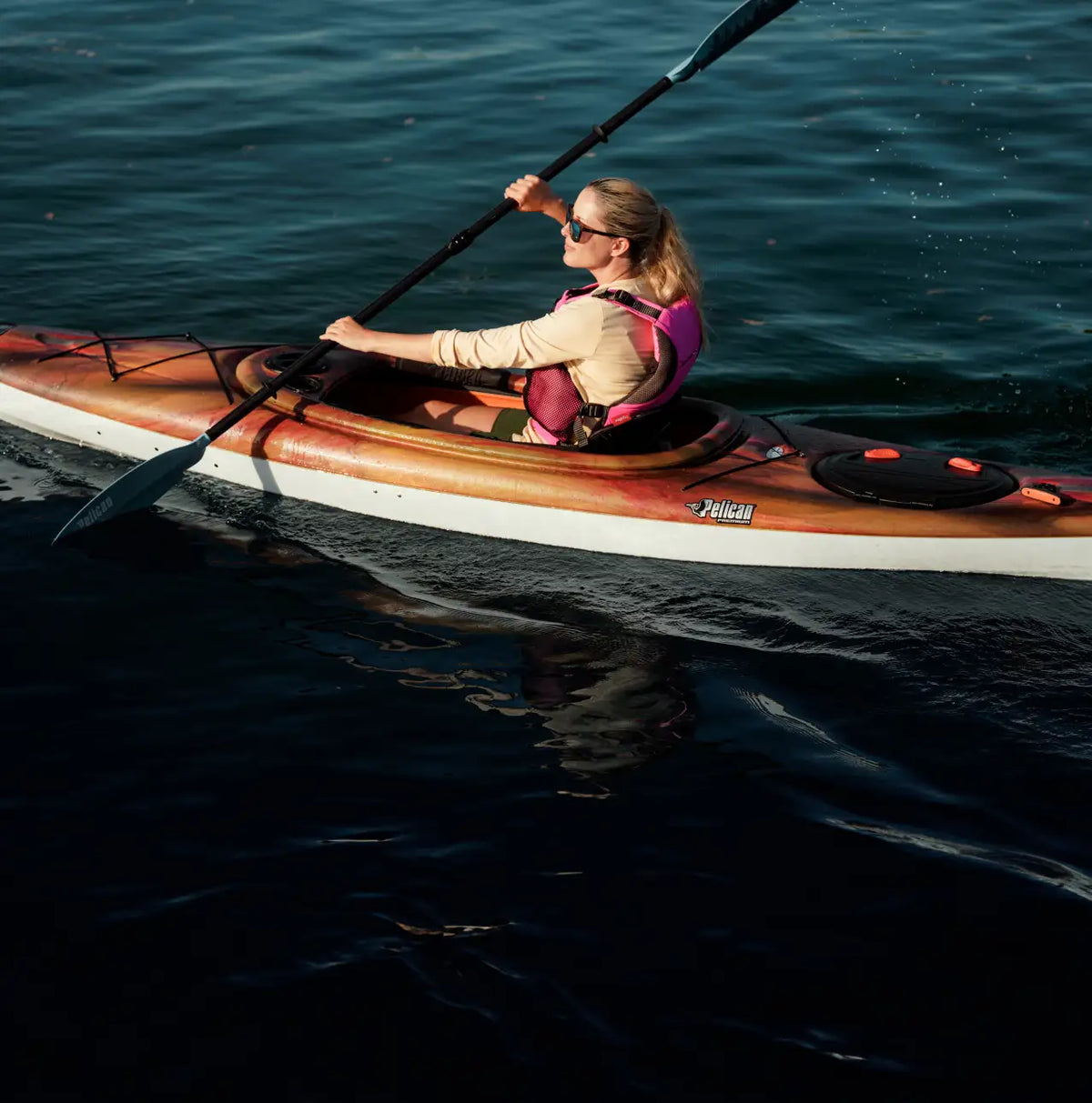 A woman paddling a Pelican Sprint Touring kayak on dark, calm water under sunlight.