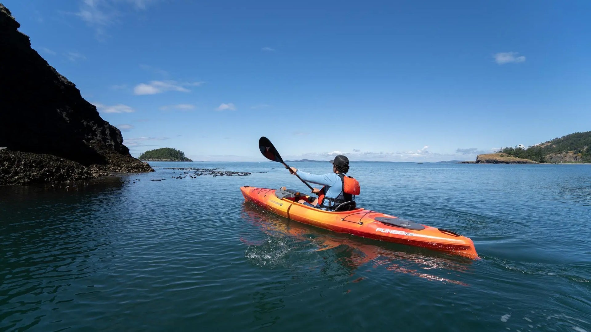 A person paddling an orange Wilderness Systems Pungo kayak on calm open water.