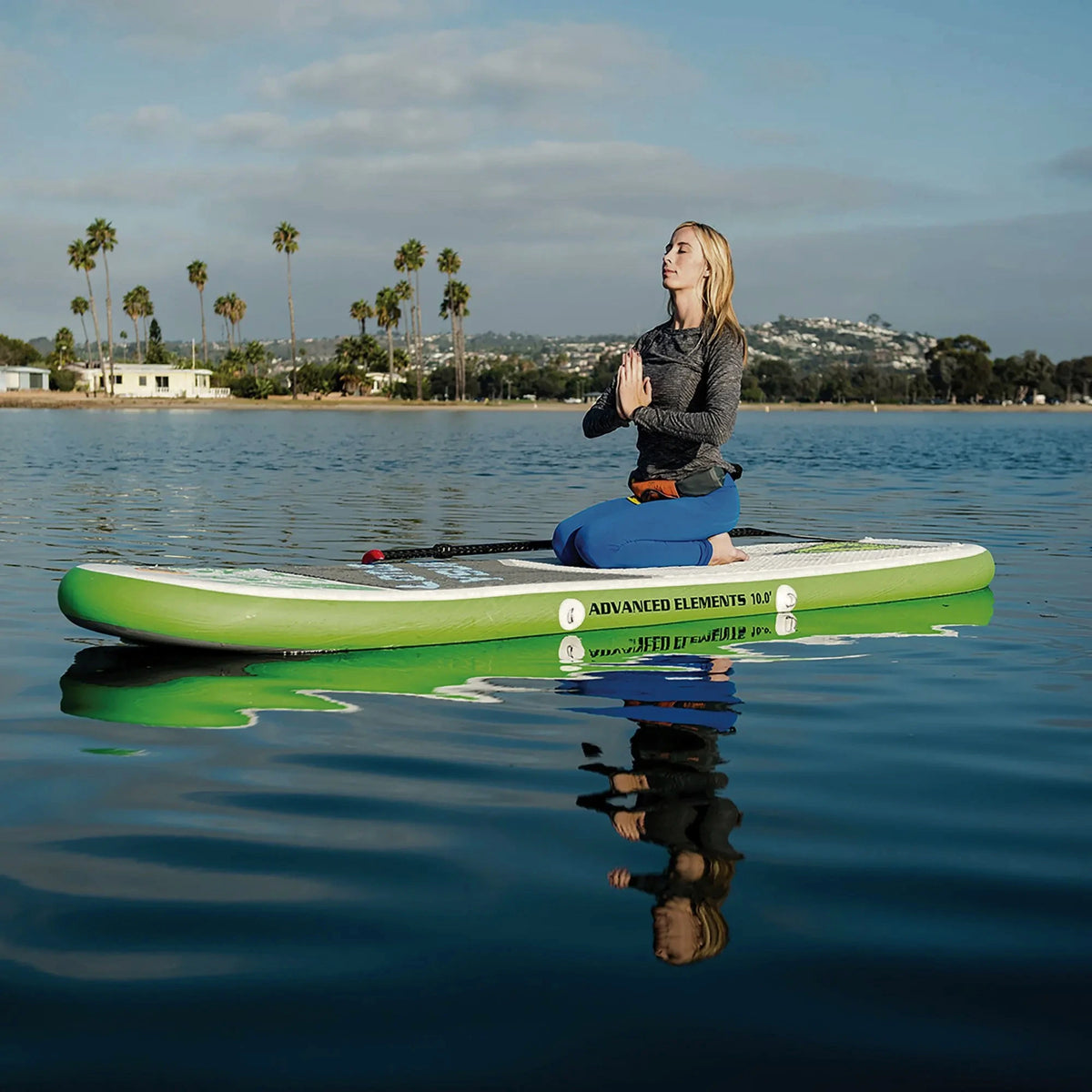 A woman kneeling in meditation pose on a green and white Advanced Elements stand-up paddleboard on calm blue water, with palm trees and hills in the background.