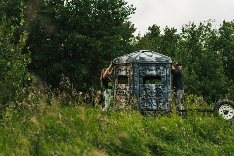 Two people setting up a camouflage Pelican CATCH hunting blind on a trailer in a field.