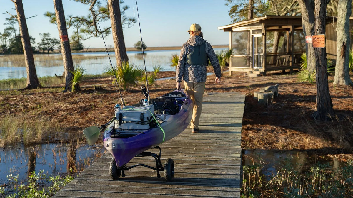 A person pulling a purple Perception Pescador Pro fishing kayak on a cart along a wooden boardwalk.