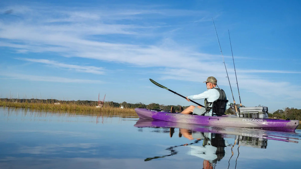 A man paddling a purple Perception Pescador Pro fishing kayak on calm water under a blue sky.