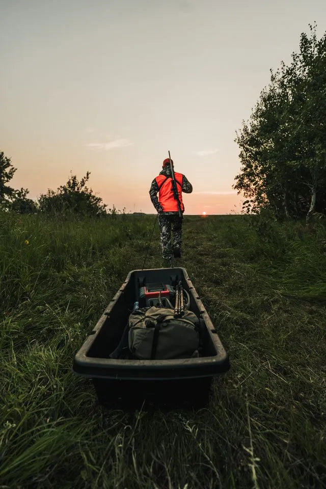 A hunter in high-visibility safety gear walking through a field at sunrise next to a Pelican CATCH utility sled.